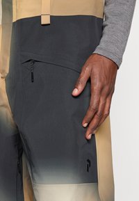 Κοντινό πλάνο ενός χεριού που rests πάνω σε παντελόνι με χρωματικούς διαχωρισμούς, το οποίο έχει τσέπη με φερμουάρ και διακριτό λογότυπο στο μηρό.