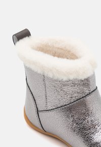 Bottine en argent métallique avec surface texturée, doublure en fausse fourrure crème et semelle en caoutchouc brun. Comprend une languette pour un enfilage facile.
