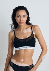 Crna čipkana bralette s prozirnim vrhom, koja ima široki bijeli pojas s natpisom "GUESS". Uz to su usklađeni crni gaćice s čipkastim rubovima.