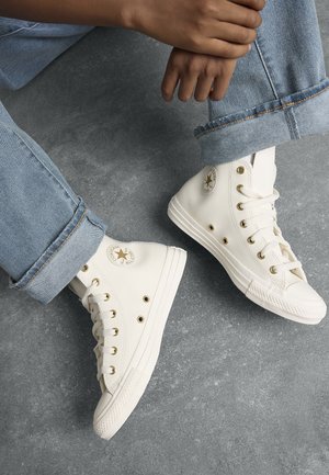 Baskets montantes blanches en cuir, avec œillets dorés, un patch logo rond et semelles en caoutchouc texturées, associées à un jean bleu clair.