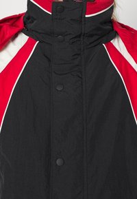 Sort vinterjakke med rød og hvid farveblokering på skuldrene, høj krave og sorte tryknapper ned langs forsiden.