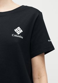 Μαύρο κοντομάνικο μπλουζάκι με λευκό λογότυπο Columbia στο στήθος και μικρότερο λογότυπο στο αριστερό μανίκι, φορεμένο από ένα άτομο με φόντο ένα απλό φόντο.