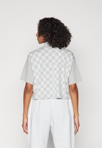 Tricou scurt, gri, cu un model de tablă de șah pe spate, mâneci scurte și o croială relaxată, realizat dintr-un material moale din bumbac.