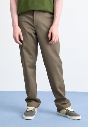 Person wearing olive green pants and two-tone gray and beige sneakers standing on a light blue floor against a white background.