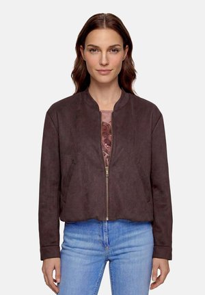 Femme aux cheveux bruns porte une veste zippée marron foncé par-dessus un haut à motifs et un jean bleu clair, debout sur un fond blanc.