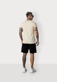 Beige short-sleeve t-shirt, black shorts, and white sneakers. Features tattoos on arms and legs, with a smooth texture.