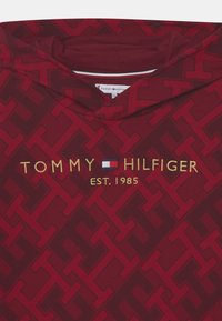 Felpa rossa con cappuccio caratterizzata da un motivo testurizzato, logo ricamato "TOMMY HILFIGER EST. 1985" in oro, con un piccolo dettaglio della bandiera in blu e bianco.
