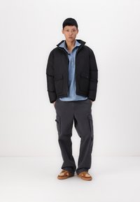 Jachetă puffer neagră cu închidere cu fermoar și două buzunare frontale, asociată cu o cămașă cu guler de culoare albastru deschis și pantaloni cargo gri. Încălțăminte maro.