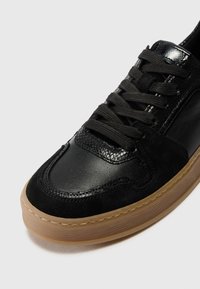 Baskets noires en cuir texturé et daim, lacets noirs et semelle en caoutchouc marron clair sur fond gris.
