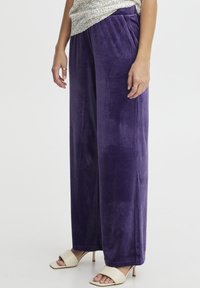 Fioletowe welurowe spodnie o szerokich nogawkach z gładką fakturą i dwiema bocznymi kieszeniami, zestawione z jasnobeżowymi sandałami na obcasie.