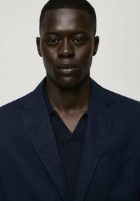 Marinblå blazer med nyckelkrage och bröstficka, lager på en texturerad mörk tröja. Slät tyg med strukturerad passform, minimal detaljering.