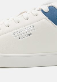 White leather sneaker with blue accent. Features embossed branding "JACK & JONES EST. 1990" on the side. Textured material, rubber sole.