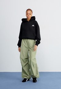 Black hoodie with a front pocket, paired with loose, light olive green pants. Model wears black sneakers. Simple, casual outfit.