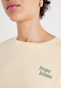 Bežové bavlněné tričko s jemnou texturou a kulatým výstřihem, zdobené zeleným logem "Pepe Jeans" na levé straně hrudi.