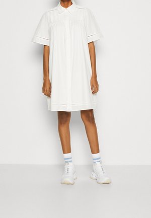 Osoba ubrana w luźną białą sukienkę z krótkim rękawem i kołnierzykiem, białe trampki oraz białe skarpetki z dwoma jasnoniebieskimi paskami, stojąca na szarej podłodze.