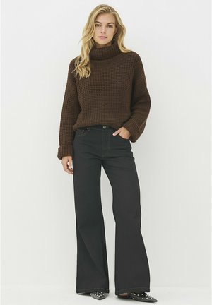 Blondinka v rjavi masivni pulover z visokim ovratnikom, črnih širokih hlačah in črnih pocinkanih špicastih čevljih, stoji ob preprostem ozadju.