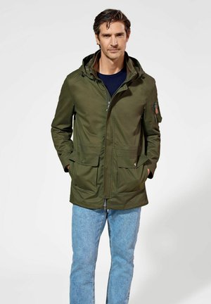 Oliwkowa parka z kapturem, z zamkiem z przodu, dwiema dużymi kieszeniami bocznymi oraz kieszonką na suwak na rękawie, zestawiona z jasnoniebieskimi dżinsami.