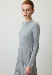 Kvinde med rødt hår i en knold iført sølvringe og en langærmet grå ribstrikket kjole foran en ensfarvet lys baggrund.