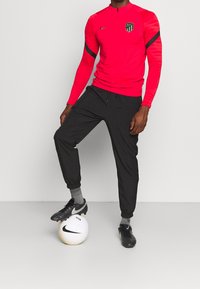Camiseta deportiva roja de manga larga con media cremallera, logo de Nike y pantalones negros. El jugador está de pie sobre un balón de fútbol blanco, usando botas negras.