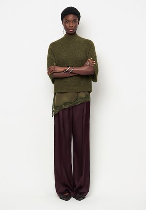 Black woman with short hair stands with arms crossed wearing green knit top, olive lace-trim layer, wide purple trousers, and silver jewelry.
