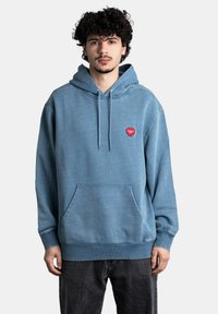Sudadera azul clara con un bolsillo frontal, capucha ajustable con cordón y un pequeño parche en forma de corazón rojo con texto. Material de mezcla de algodón.
