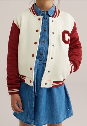 Personne portant une veste varsity crème et rouge avec un «C» sur la poitrine, par-dessus une robe en denim bleu boutonnée, main détendue sur le côté.