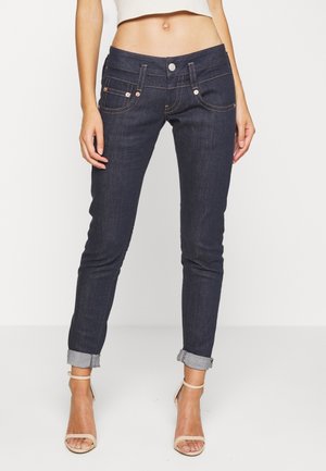 Woman wearing dark blue rolled-up skinny jeans and beige high-heeled sandals, standing against a plain light background.