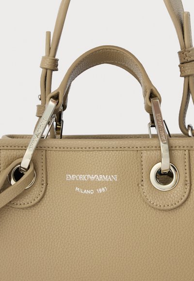 Τσάντα χειρός από μπεζ δέρμα με διπλές λαβές, ασημένια μεταλλικά στοιχεία και ανάγλυφο λογότυπο. Υφή σε φινίρισμα και κυκλικά μεταλλικά διακοσμητικά.