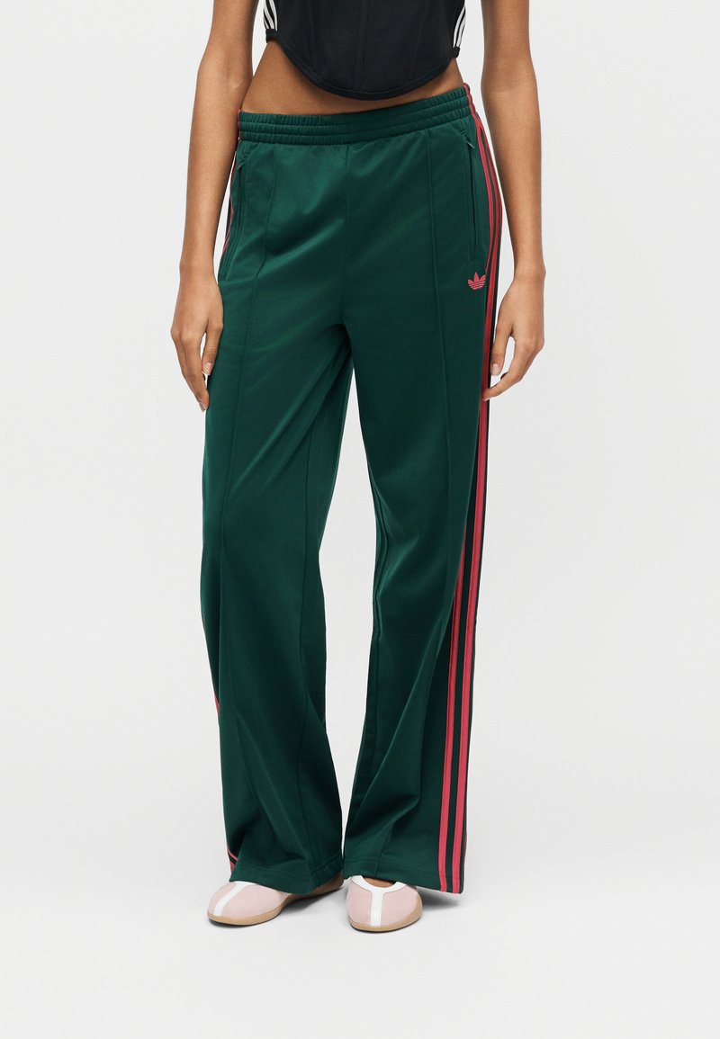 Personne portant un pantalon de survêtement large vert foncé avec des bandes latérales roses et un petit logo rose, associé à des chaussures rose pâle.