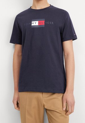 Man draagt een marineblauw Tommy Hilfiger T-shirt met logo op de borst en beige broek, staand tegen een witte achtergrond.