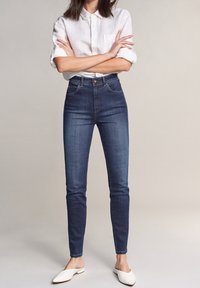 Jeans de tiro alto de color azul oscuro con un corte ajustado, diseño clásico de cinco bolsillos y detalles de costura sutil, combinados con una camisa blanca de botones.