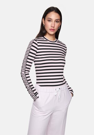 Frau mit einem rosa und schwarzen, horizontal gestreiften Langarmshirt und hellrosa Jogginghose mit Kordelzug, die mit Händen in den Taschen steht.