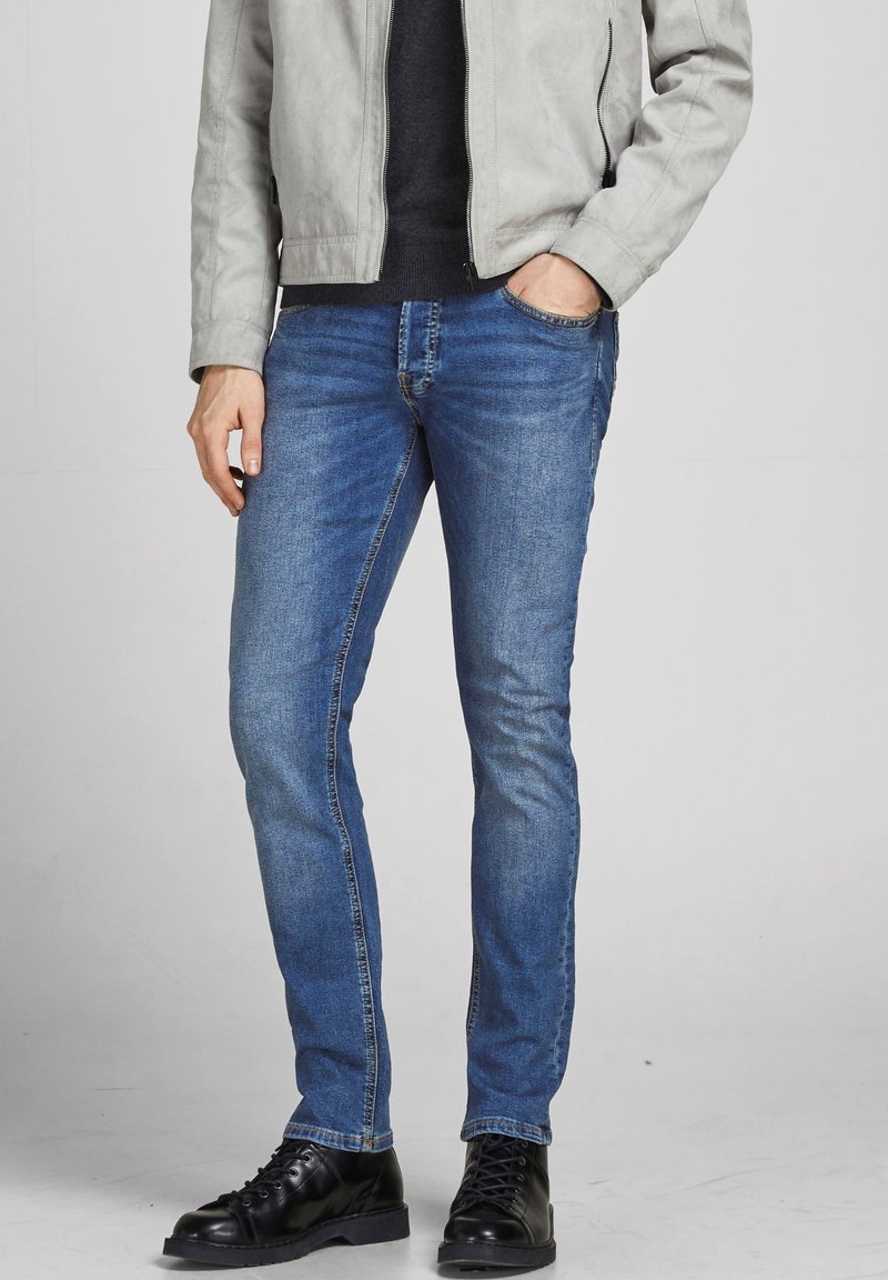 Slim-fit blauwe jeans van denim met een lichte vervaging, gecombineerd met een lichtgrijze jas en zwarte veterschoenen.