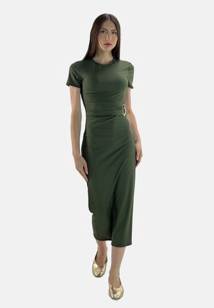Grünes, figurbetontes Kleid aus glattem Stoff, knielang mit kurzen Ärmeln, mit einem seitlichen Schnallenakzent und gerader Silhouette, kombiniert mit goldenen Schuhen.