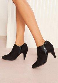 Botas de tobillo de ante negro con punta afilada, detalles brillantes en charol negro, cremallera lateral y un tacón de 3 pulgadas. Textura suave en general.