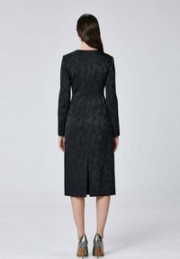 Robe noire à manches longues, motif floral, taille ajustée et fente au dos. Des chaussures à talons hauts argentées complètent la tenue.