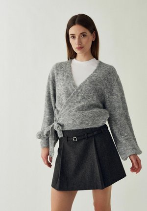 Femme debout devant un fond clair portant un pull gris croisé sur un haut blanc et une jupe plissée foncée avec une fine ceinture.