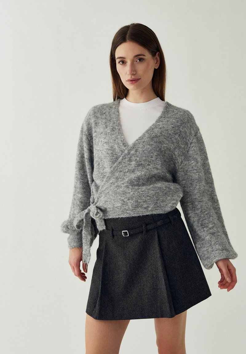 Femme debout devant un fond clair portant un pull gris croisé sur un haut blanc et une jupe plissée foncée avec une fine ceinture.