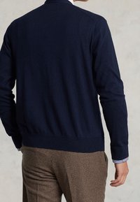 Mörkblå stickad cardigan med långa ärmar, ribbad mudd och nederkant. Visas från baksidan med rutiga bruna byxor. Slät textur.