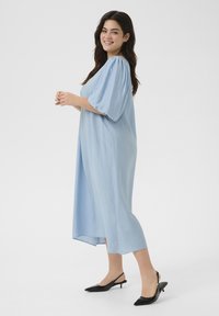 Robe bleu clair, jusqu'au genou, avec des manches bouffantes et une coupe ample. Associée à des escarpins noirs à talon slingback, avec un design à bout pointu.
