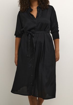 Kobieta w czarnej, zapinanej na guziki sukience midi z rękawami do łokcia i wiązanym paskiem w talii, stojąca na tle jednolitego jasnego tła.