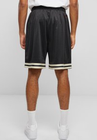 Svarte atletiske shorts med mesh-struktur, elastisk livlinje og kremfargede og svarte stripes detaljer nederst. Bakfra, som viser ben og joggesko.