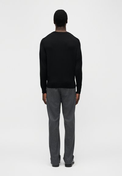 Homme debout de dos, portant une chemise noire à manches longues et un pantalon gris coupe droite, sur un fond blanc uni.