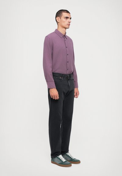 Paarse langharige shirt met knoopsluiting, zwarte jeans en groene suède sneakers met witte veters, staande tegen een effen achtergrond.