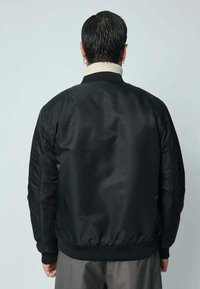 Veste bomber noire avec une surface texturée, des poignets et un col côtelés, et une coupe décontractée. Présente des détails de couture minimalistes sur les manches.