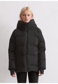 Blonde Frau in einer schwarzen Daunenjacke mit hohem Kragen und Reißverschlusstaschen, die vor einer schlichten weißen Wand steht.