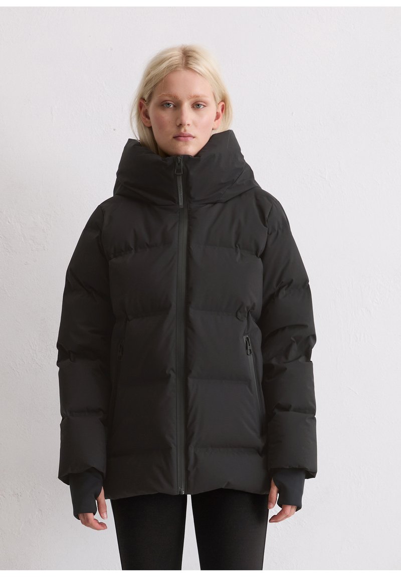 Blonde Frau in einer schwarzen Daunenjacke mit hohem Kragen und Reißverschlusstaschen, die vor einer schlichten weißen Wand steht.