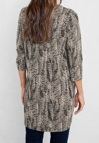 Långärmad tunika i svart och beige, med ett bladigt mönster. Tillverkad av textil med struktur och rak passform, som slutar vid mitten av låret, parat med mörka jeans.