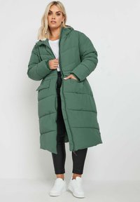 Grön vadderad lång kappa med huva, med ett quiltat mönster, sidofickor och tryckknappar, bärs över svarta byxor och vita skor.