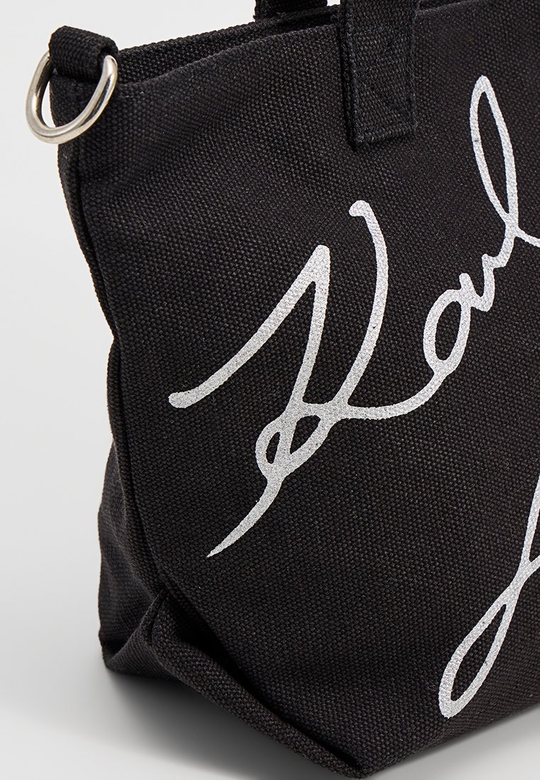 Sac fourre-tout noir en toile avec un design de signature argenté en évidence, forme structurée, doubles poignées supérieures et accents en métal.